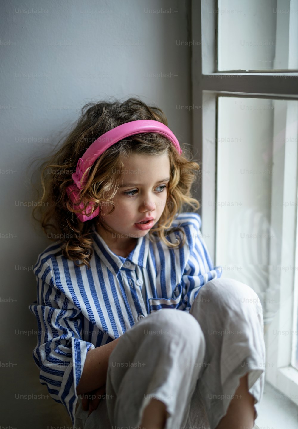 Porträt eines traurigen kleinen Mädchens mit Kopfhörern drinnen zu Hause, das auf der Fensterbank sitzt.