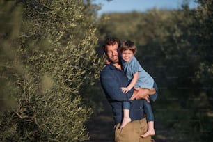 Ritratto di padre che tiene in braccio la figlia piccola, in piedi all'aperto vicino all'ulivo.
