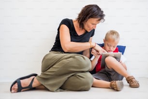 A portrait of down-syndrome school boy sitting on the floor with teacher, using tablet.