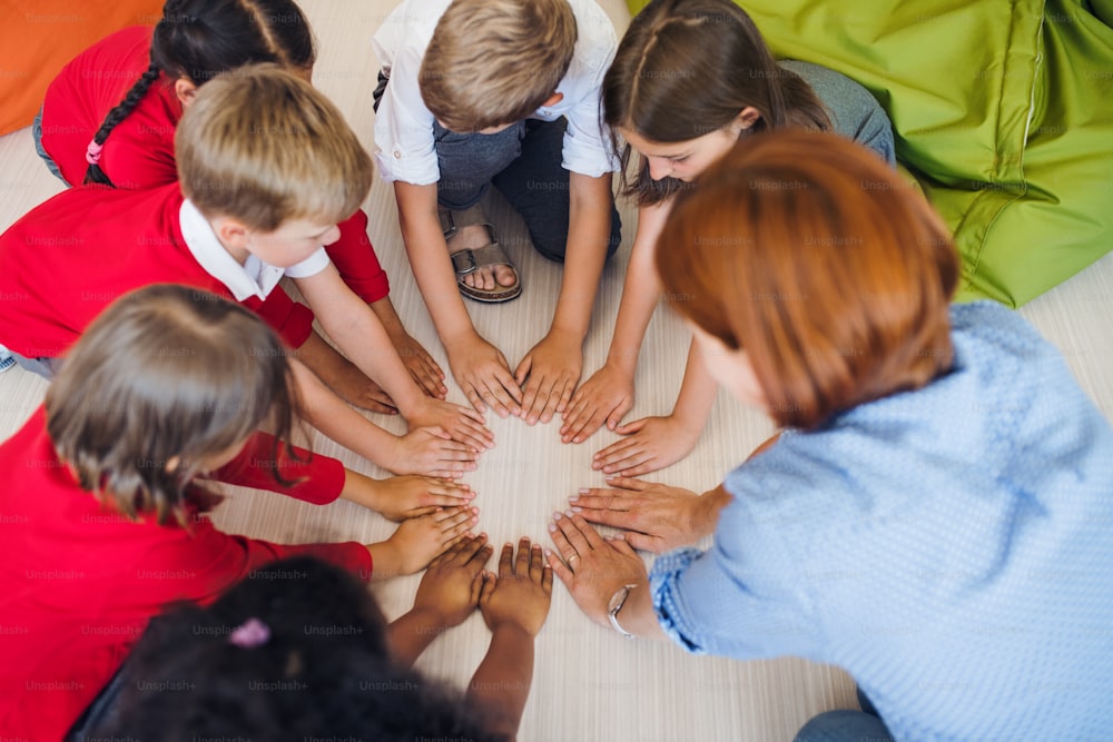 A top view of group of small school kids with teacher sitting on the floor in class, putting hands in the middle.