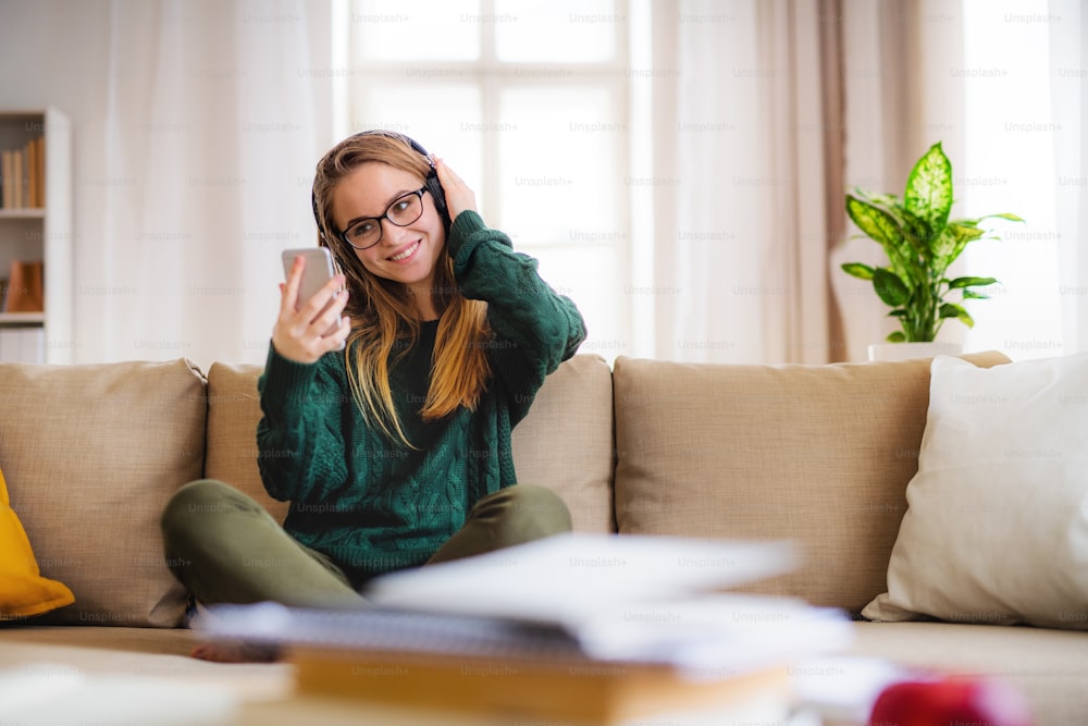 Eine junge, glückliche Studentin, die zu Hause auf dem Sofa sitzt und beim Lernen Kopfhörer benutzt.