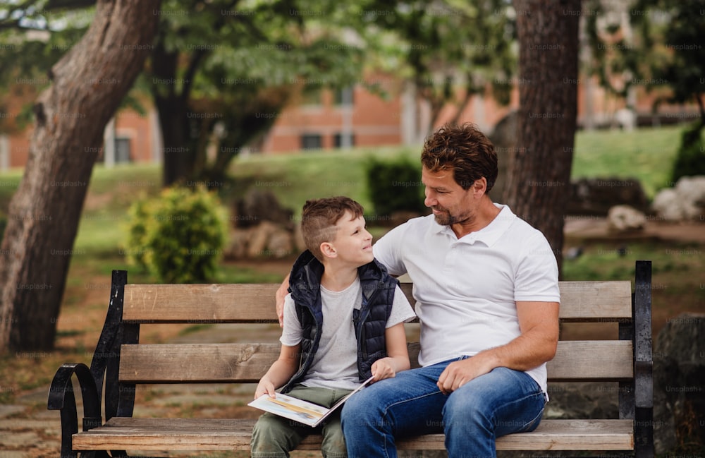 Vater mit Sohn sitzt draußen im Park in der Stadt auf der Bank und liest ein Buch.