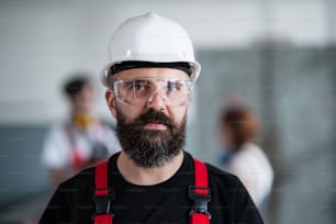 A portrait of worker with helmet and protective glasses indoors in factory looking at camera.