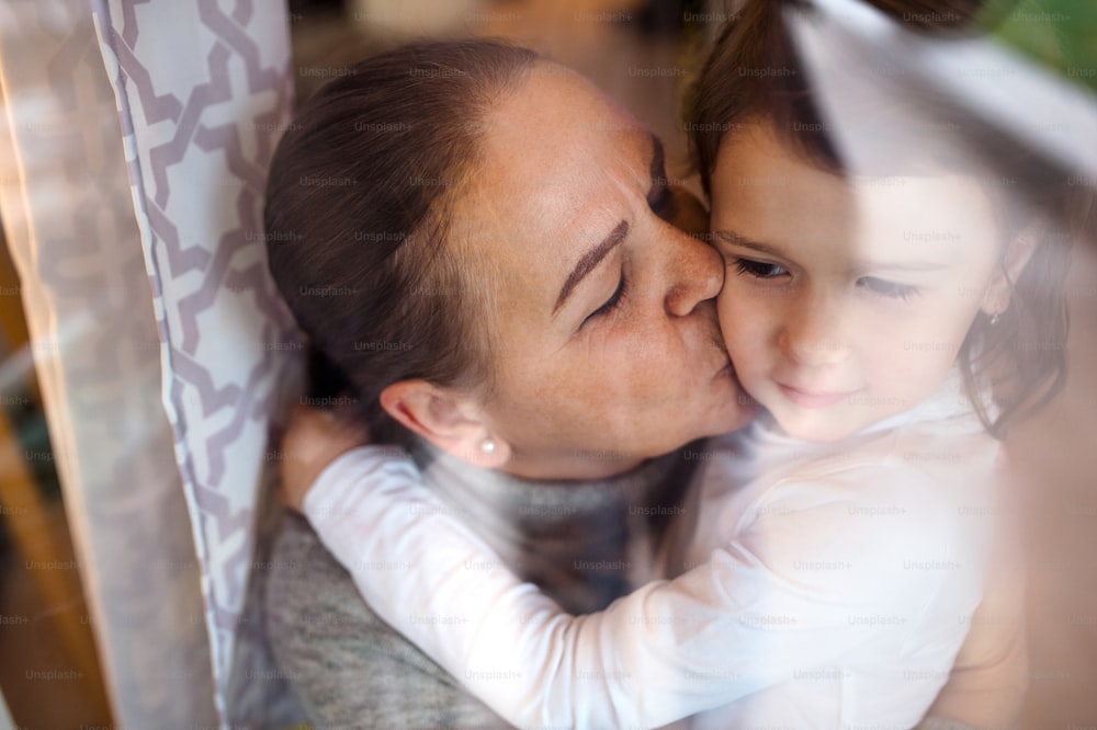 Senior grandmother kissing small granddaughter indoors at home. Shot through glass.