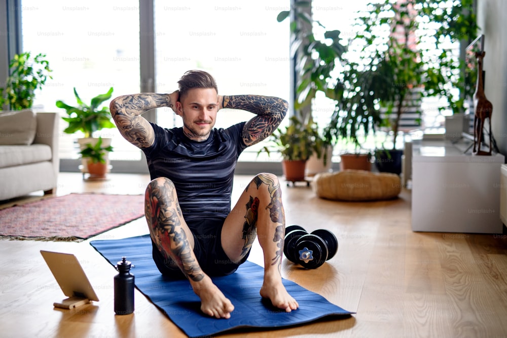 Front view portrait of young man with tablet doing workout exercise indoors at home.