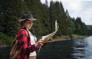 A side view of young woman standing by lake outdoors in summer nature, using map.