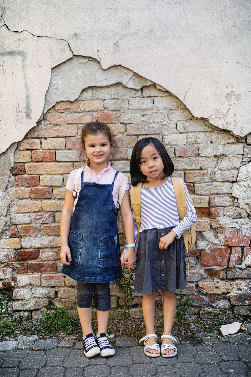 Portrait of small girls looking at camera outdoors in town, standing against old brick wall.