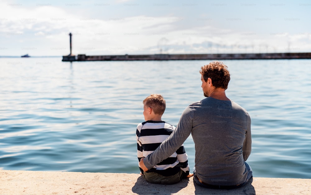 A rear view of father with small son sittting outdoors in town by the sea, talking.