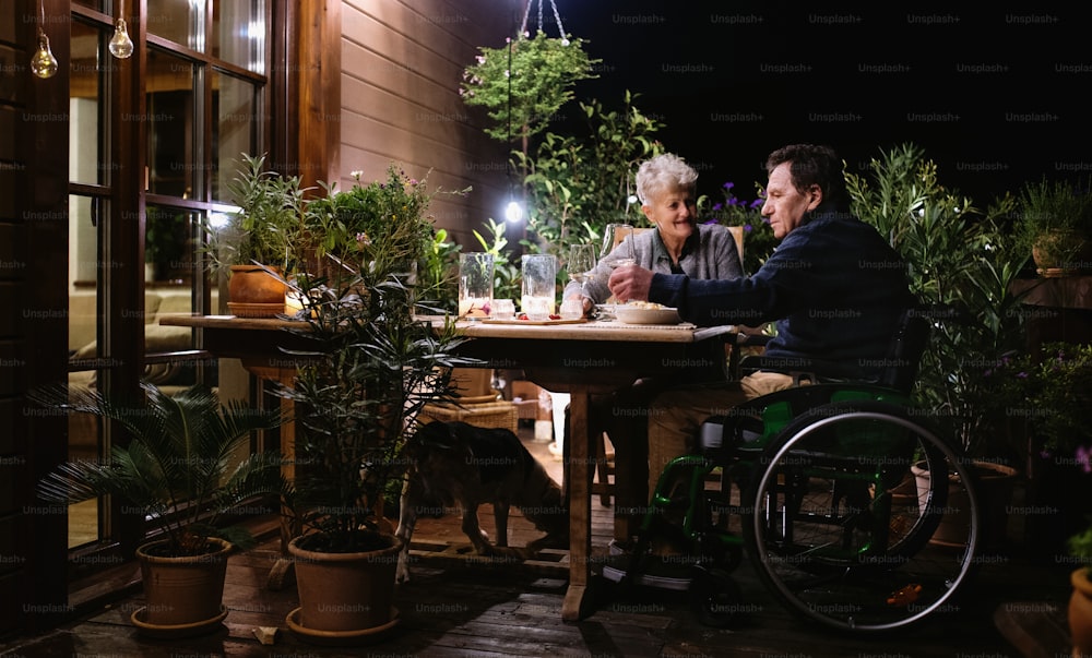 Casal idoso feliz em cadeira de rodas jantando à noite no terraço, bebendo vinho.