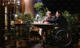Happy senior couple in wheelchair having dinner in the evening on terrace, drinking wine.