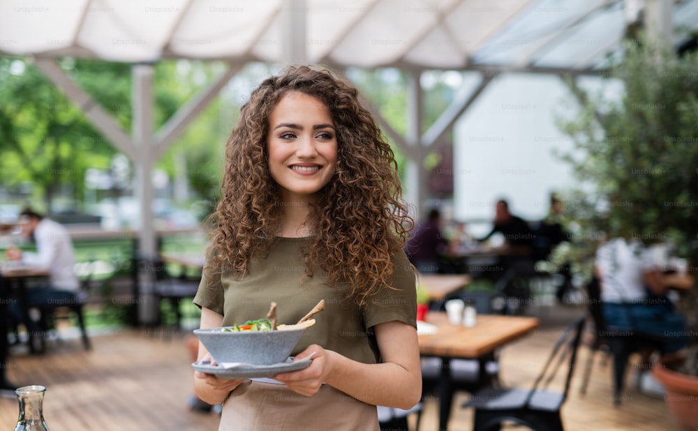 Portrait of waitress with plate standing on terrace restaurant, looking at camera.