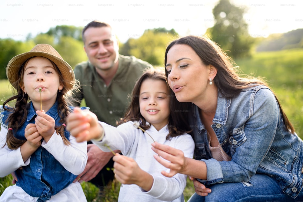 Vue de face d’une famille heureuse avec deux petites filles assises à l’extérieur dans la nature printanière, soufflant des graines de pissenlit.
