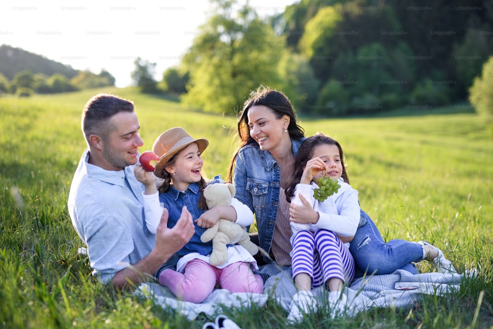 Front view of happy family with two small daughters sitting outdoors in spring nature, having picnic.