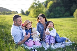 Vue de face d’une famille heureuse avec deux petites filles assises à l’extérieur dans la nature printanière, en train de pique-.
