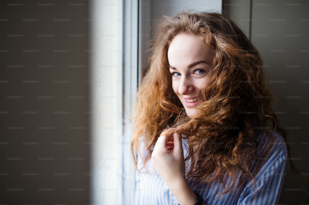 Close-up portrait of young woman standing by window indoors at home. Copy space.