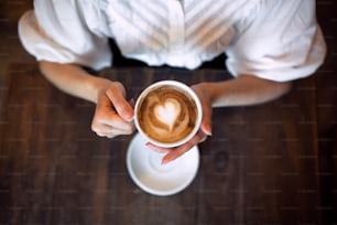 Top view of unrecognizable woman sitting at the table indoors in cafe, holding coffee.