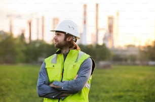 Portrait of young engineer standing outdoors by oil refinery, arms crossed.