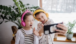 Two cheerful small girls sisters with headphones indoors at home, grimacing when taking selfie.