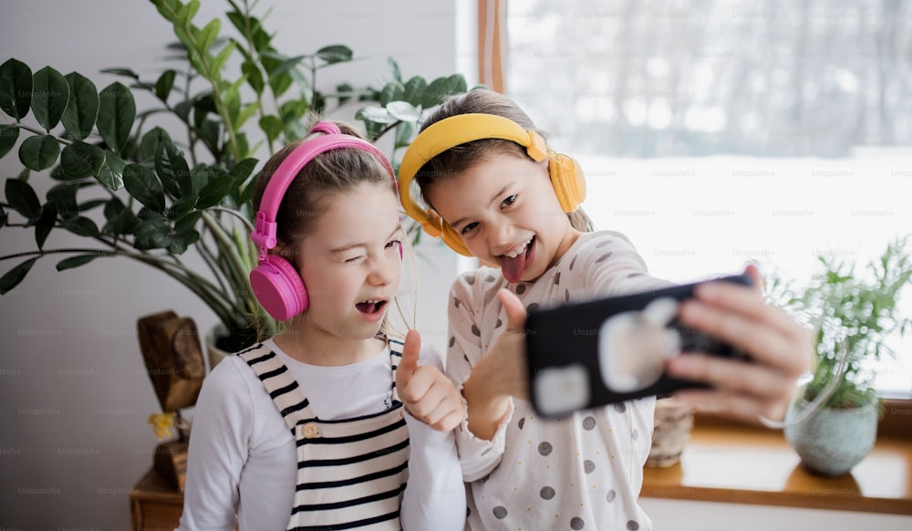 Two cheerful small girls sisters with headphones indoors at home, grimacing when taking selfie.