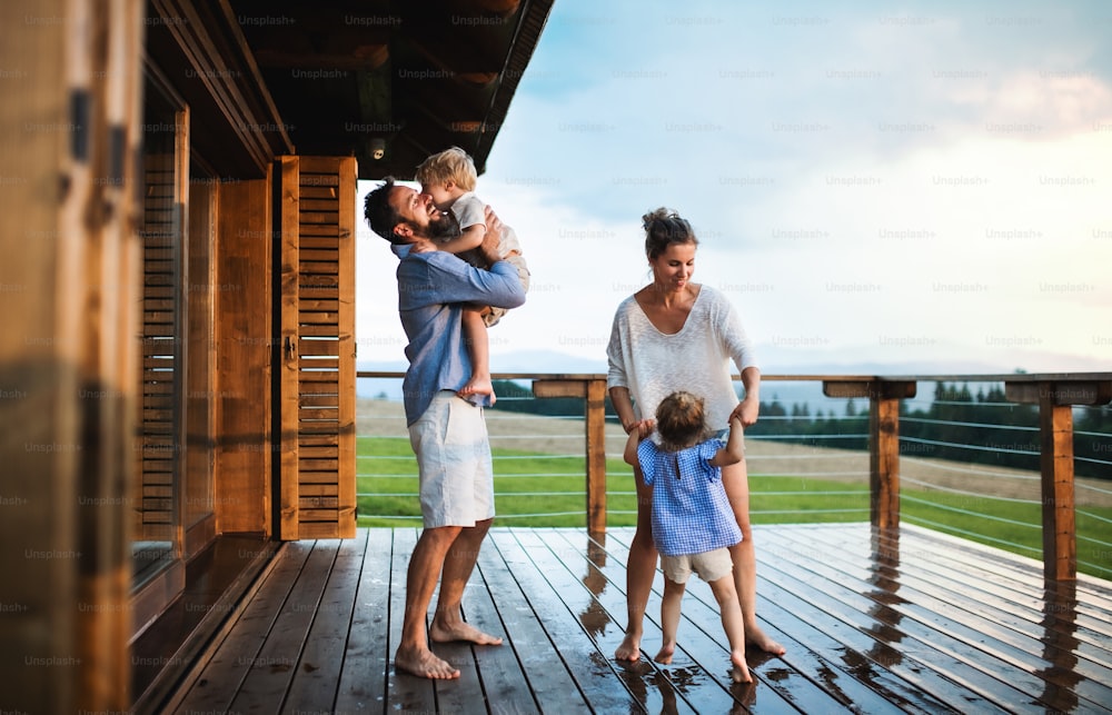 Family with happy small children playing in rain on patio by wooden cabin, holiday in nature concept.