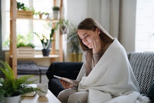 Mujer joven preocupada envuelta en una manta usando una tableta en casa, concepto de coronavirus.