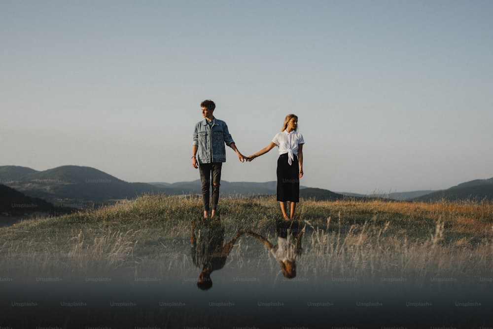 Ein Porträt eines jungen Paares, das in der Natur auf dem Land steht, Händchen haltend, aber voneinander wegschauend.