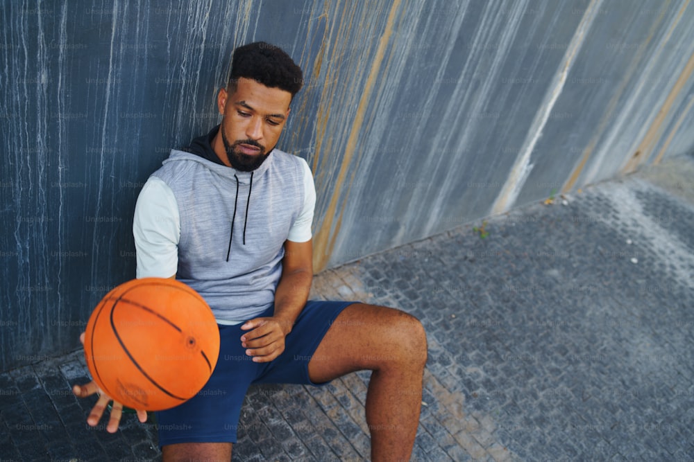 Ein junger Mann mit Basketball trainiert draußen in der Stadt, sitzt und ruht sich aus.