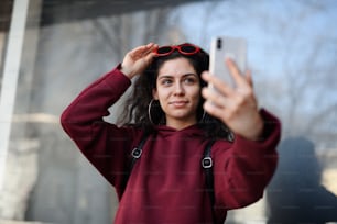 スマートフォンを持つ若い女性のポートレートが街の通りに屋外に立ち、自分撮りをする。