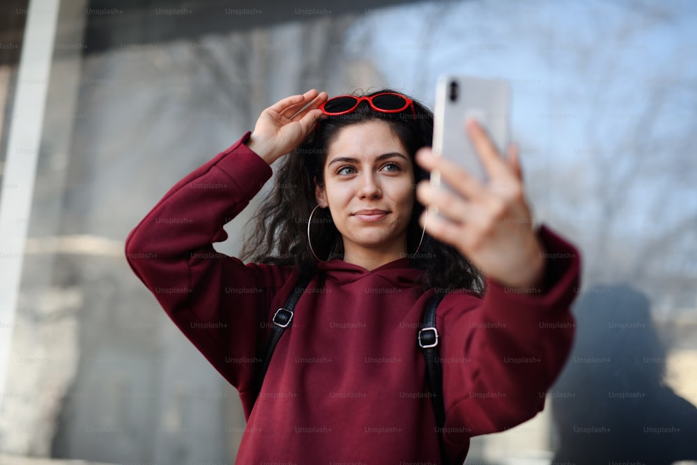 A portrait of young woman with smartphone standing outdoors on street in city, taking selfie.