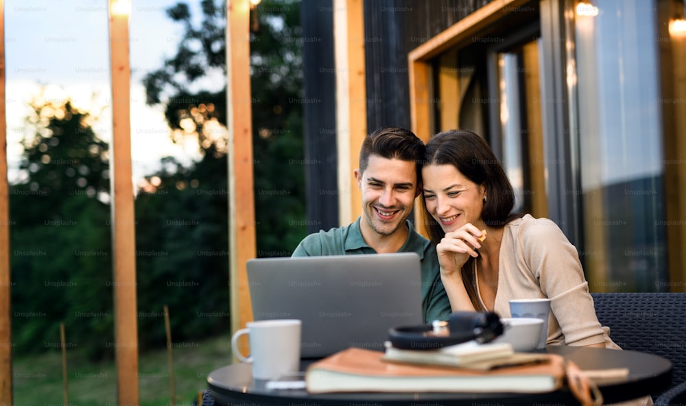 Jovem casal com laptop sentado ao ar livre, fim de semana em casa de contêiner no campo.