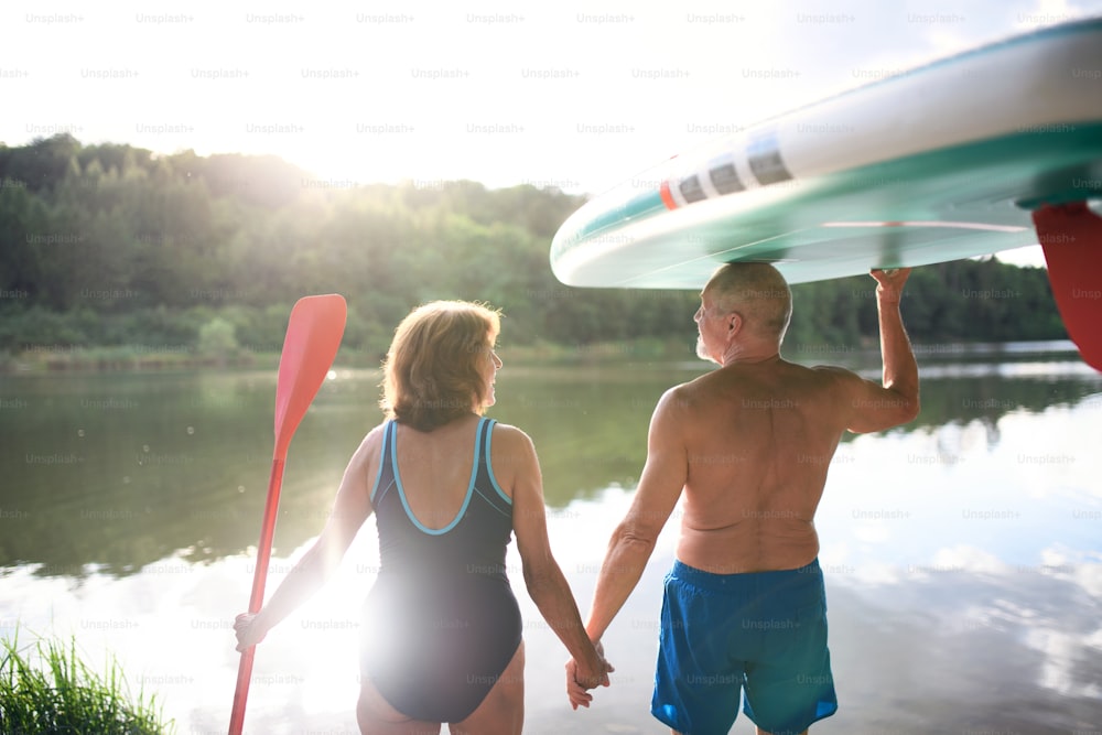 Rear view of senior couple carrying paddleboard by lake in summer, talking.