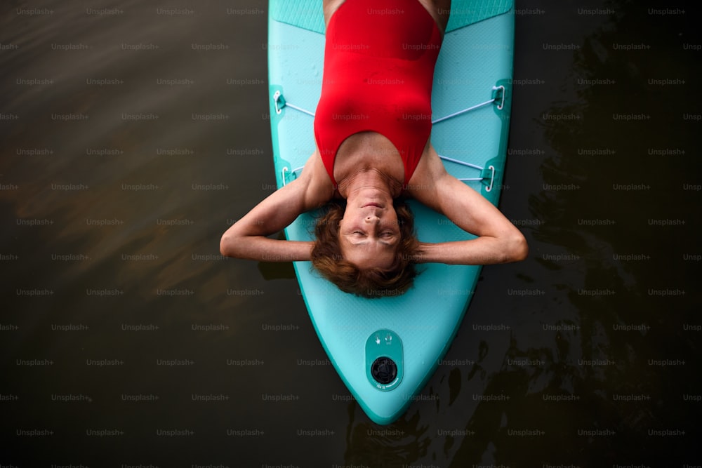 Top view of contented senior woman lying on paddleboard on lake in summer, resting.