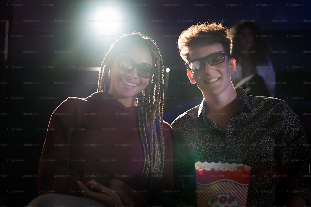 Young couple with 3d glasses and popcorn watching movie in cinema, looking at camera.