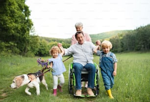 車椅子に乗った高齢の祖父母と犬を連れて、自然の中の牧草地を散歩する小さな子供たち。