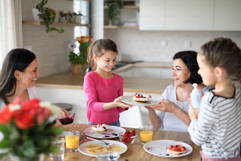 Des petites filles heureuses avec leur mère et leur grand-mère qui mangent des crêpes à l’intérieur à la maison, qui déjeunent.