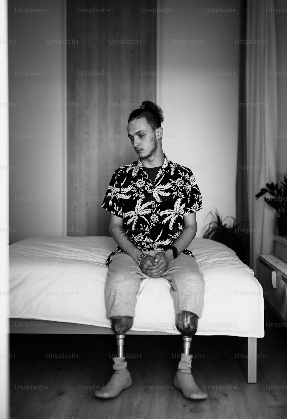 A black and white portrait of disabled young man sitting in bed indoors at home, leg prosthetic concept.