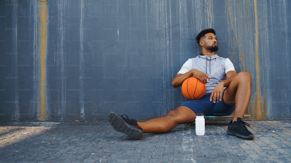 Un jeune homme avec un ballon de basket-ball faisant de l’exercice à l’extérieur en ville, assis et se reposant.