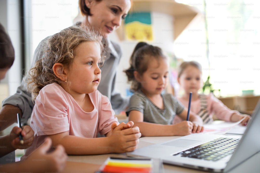 Eine Gruppe kleiner Kindergartenkinder mit Lehrer drinnen im Klassenzimmer, mit Laptop.