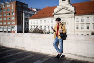 A portrait of young man standing outdoors in city, using smartphone.