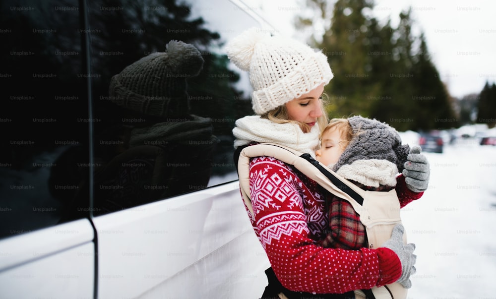 Portrait of mother with sleeping small daughter in carrier standing by car in winter nature.