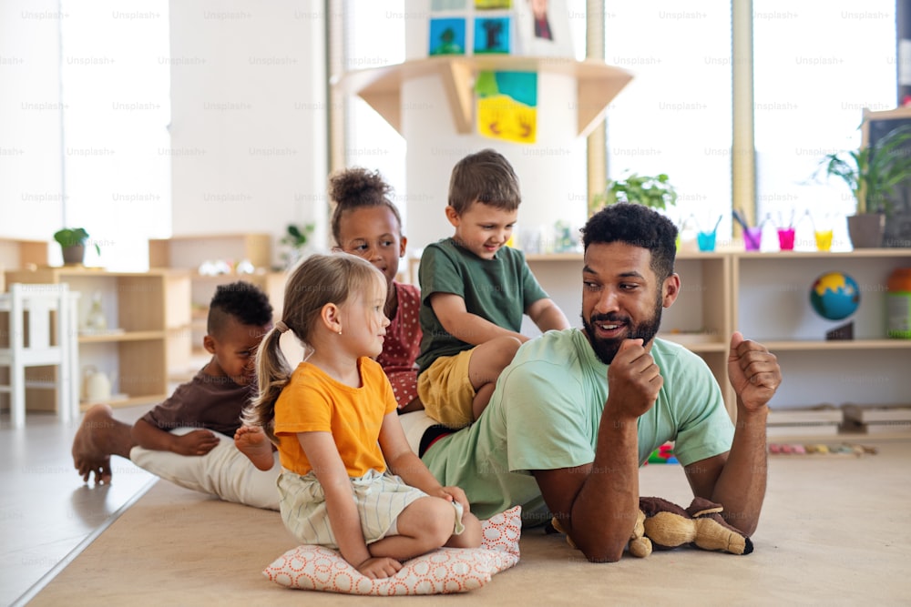 Eine Gruppe kleiner Kindergartenkinder mit einem männlichen Lehrer, der drinnen im Klassenzimmer auf dem Boden sitzt und spielt.