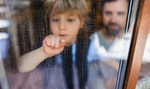 A sad boy with unrecognizable father looking through dirty window, housework concept.