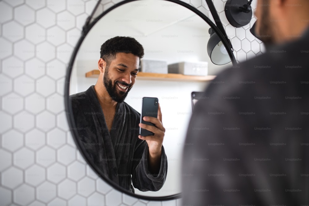 A young man with coffee and bathrobe indoors in bathroom at home, taking selfie.