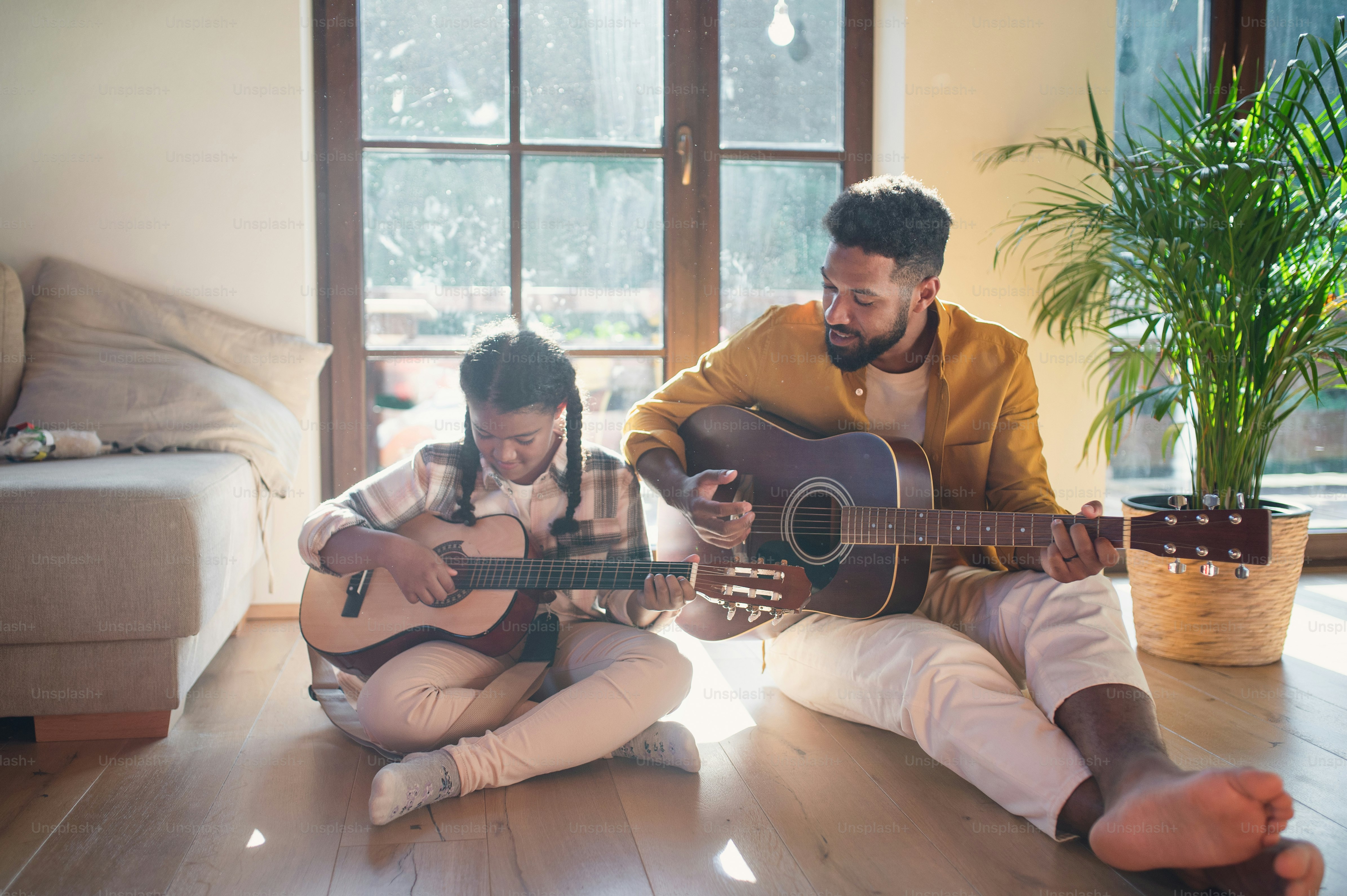 A front view of happy father with small daughter indoors at home, playing guitar.
