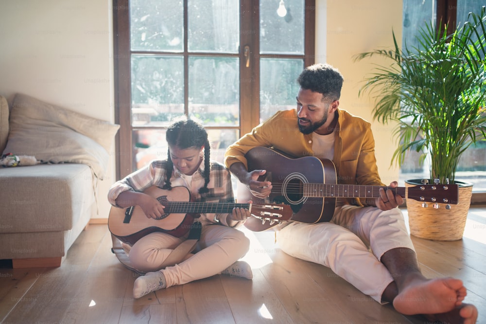 Una vista frontal de un padre feliz con una hija pequeña en el interior de la casa, tocando la guitarra.