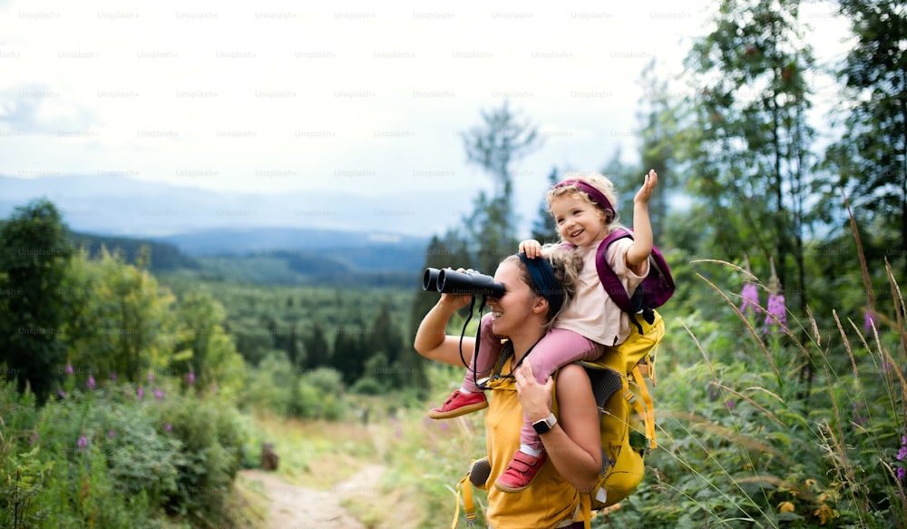 Side view of mother with small toddler daughter hiking outdoors in summer nature, using binoculars.