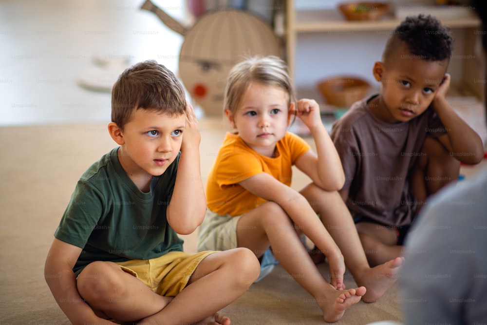 A group of small nursery school children sitting on floor indoors in classroom, listening to teacher.