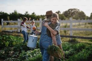 Une agricultrice adulte heureuse étreignant son camarade âgé tenant un panier avec des légumes cultivés à la maison à l’extérieur de la ferme communautaire.