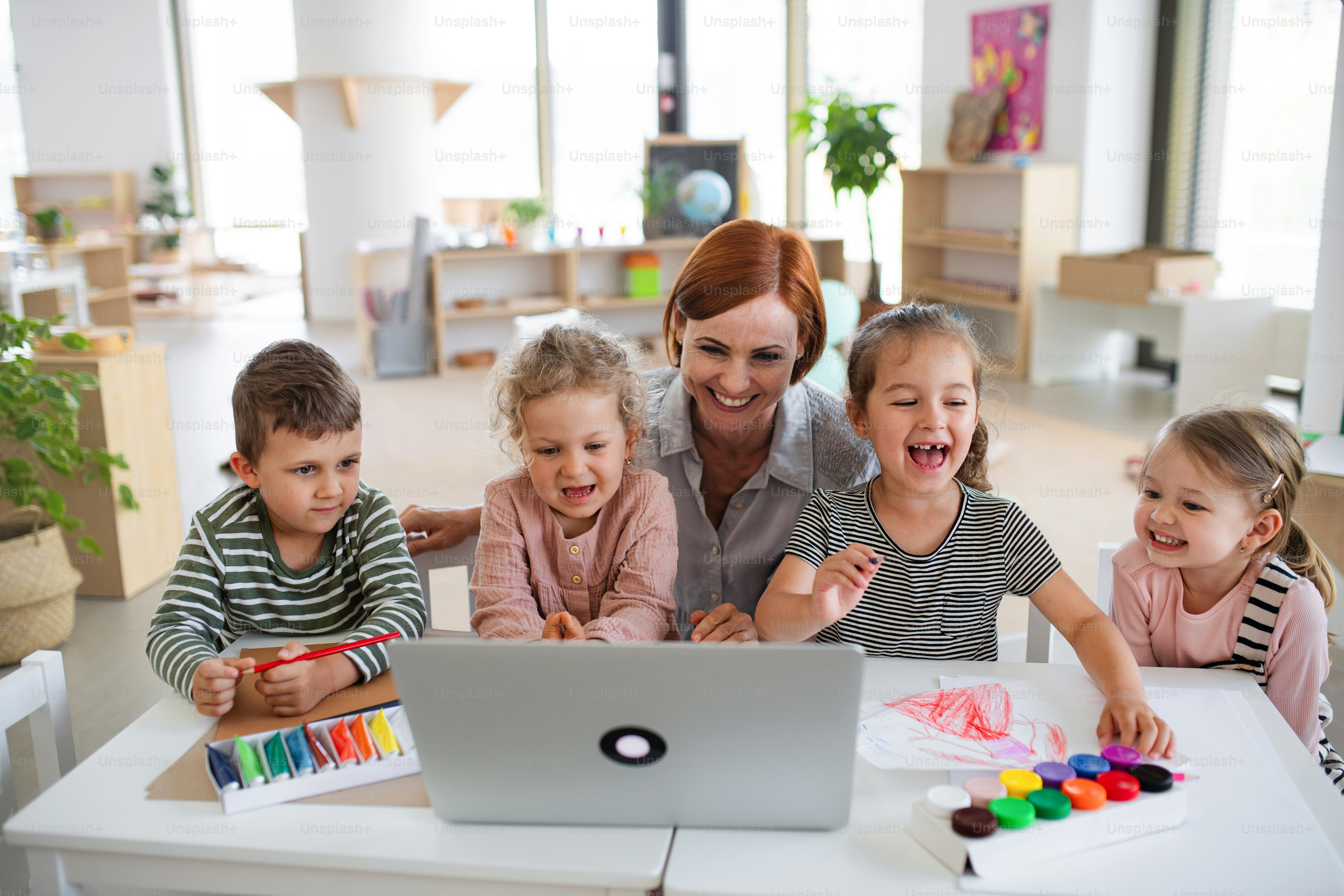Un grupo de pequeños niños de guardería con maestra en el interior del aula, usando una computadora portátil