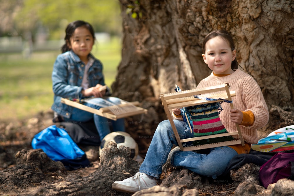 Retrato de niños pequeños con telar de mano sentados al aire libre en el parque de la ciudad, concepto de educación grupal de aprendizaje.